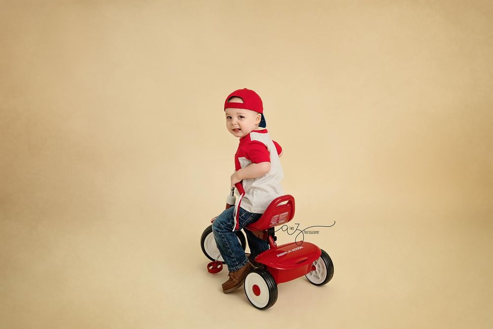 Child in red cap riding classic Radio Flyer tricycle against neutral backdrop.