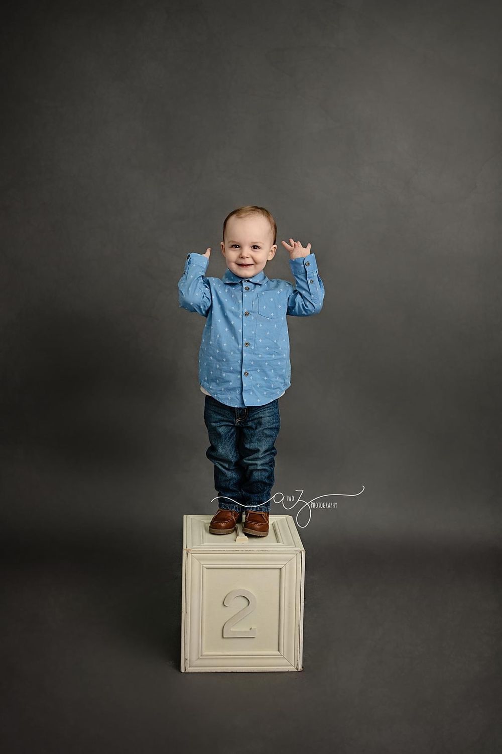 Child in denim shirt standing on numbered block against grey studio backdrop.