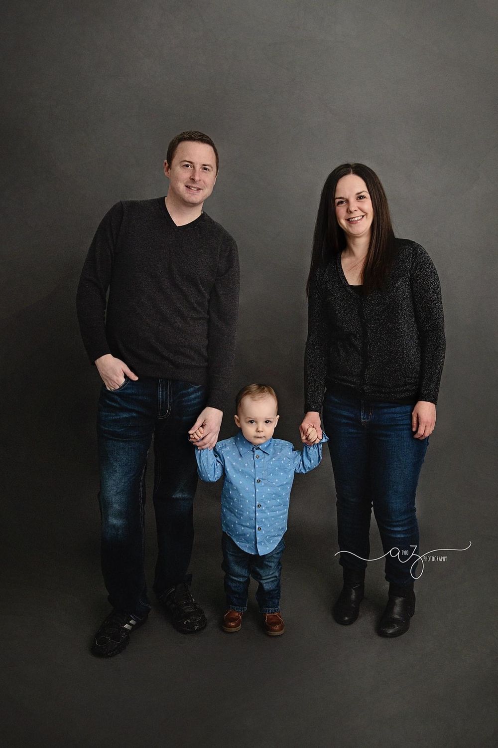 A family of three poses together in casual attire against a grey studio background.