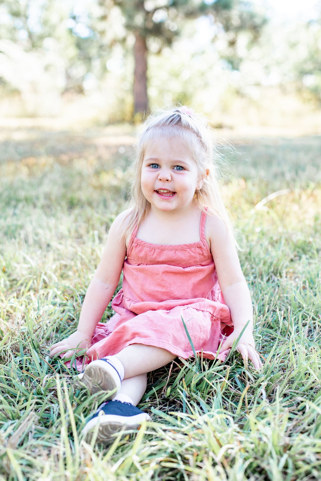 Toddler girl sits in the grass and smiles in Black Forest, Colorado.