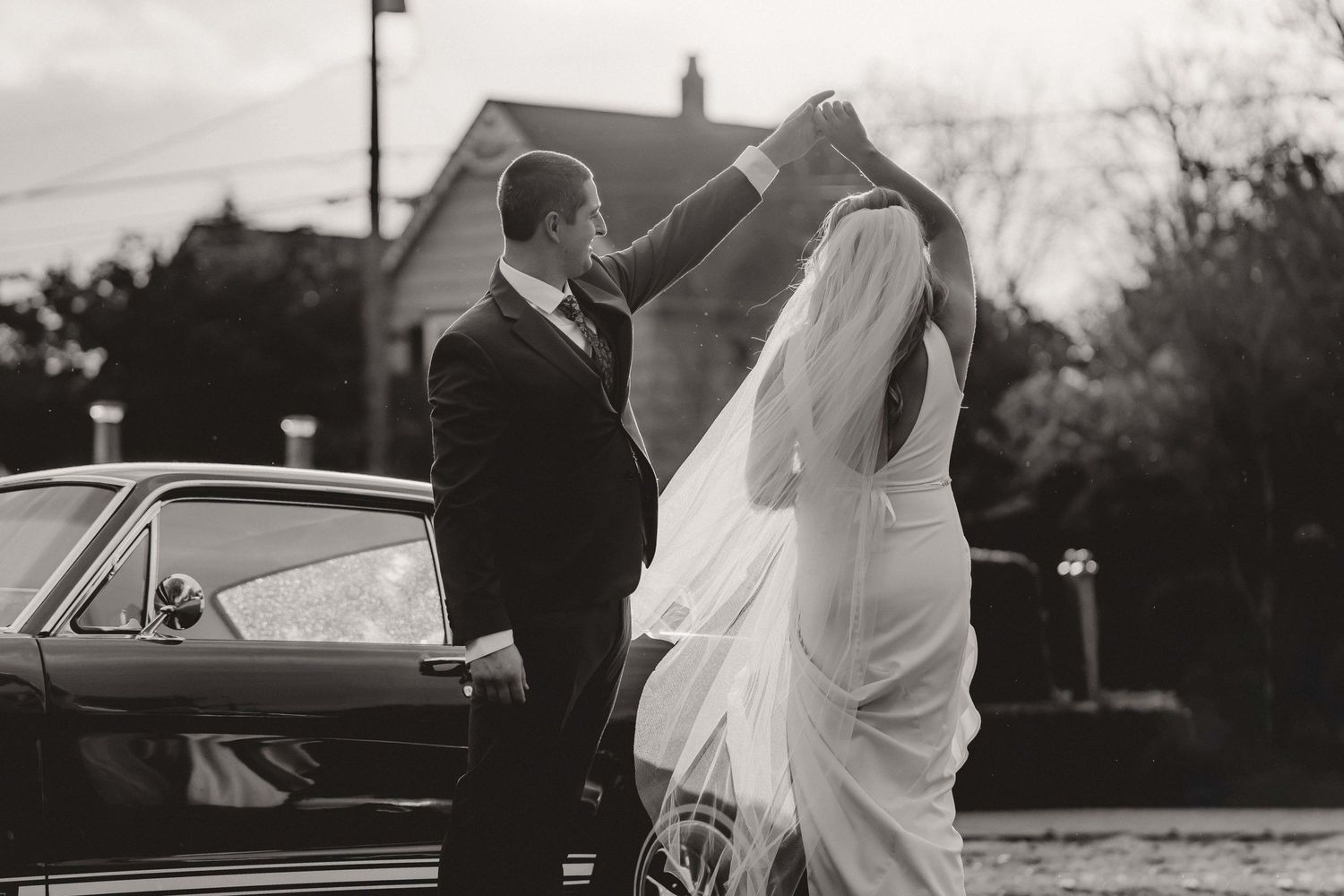 Bride and groom dancing on the street with a vintage car in black and white.