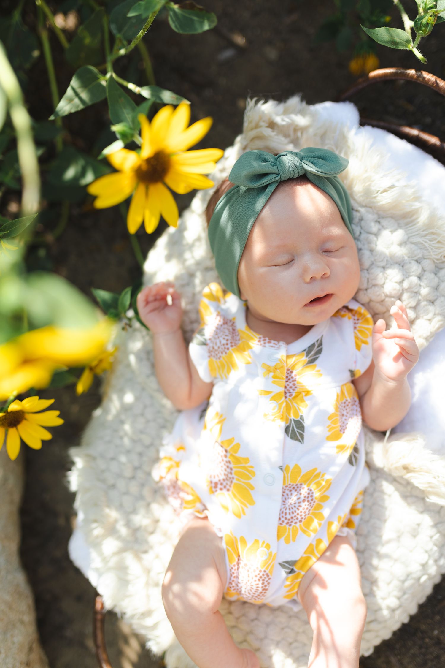 Baby in sunflower print outfit with green bow headband rests on white blanket among yellow flowers.