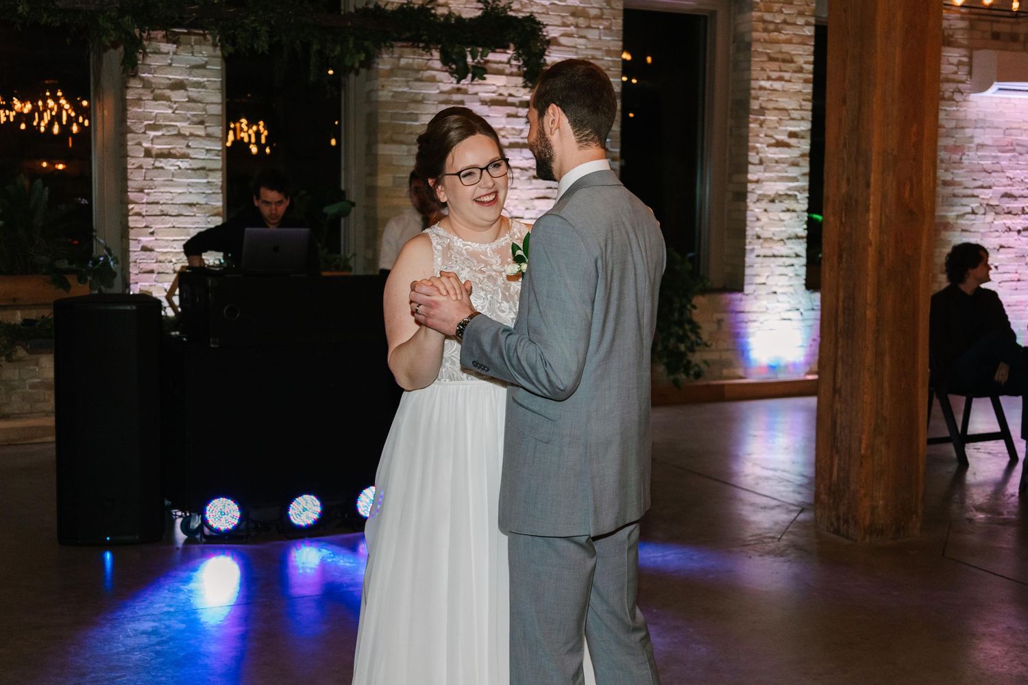 A couple shares their first dance in a rustic brick-walled venue with blue lighting.