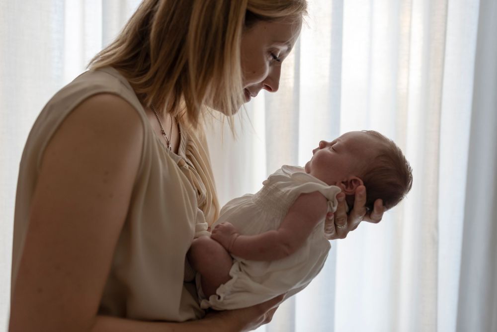 A tender moment of a blonde mother in a cream dress gently holding her newborn baby by a bright window.