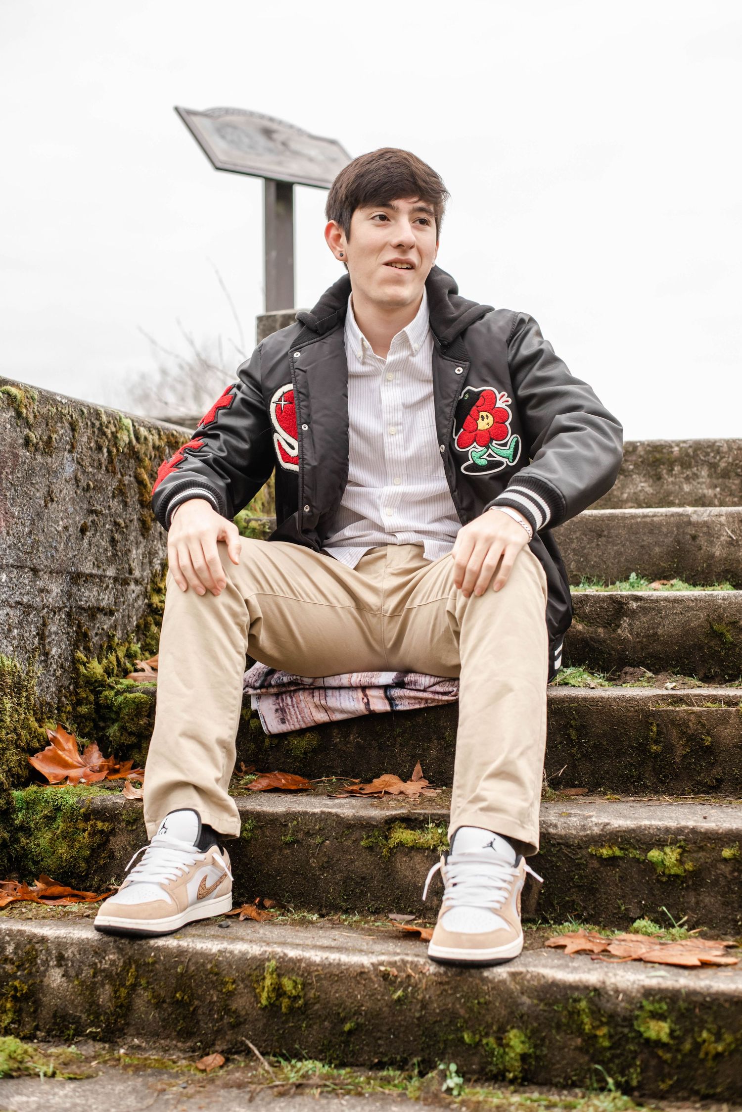 Person in black jacket and khakis sits casually on concrete steps outdoors.