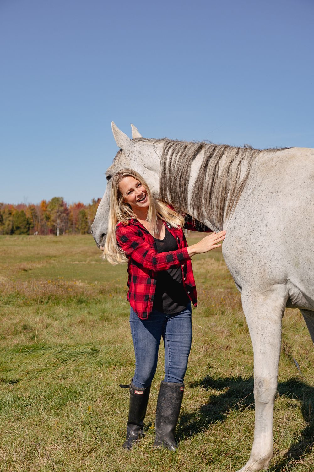 A person wearing a red plaid shirt and jeans hugs a white horse in a sunny pasture.