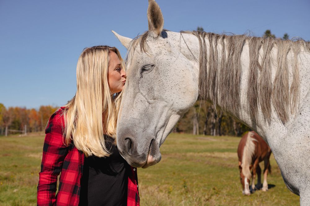 A person in a red plaid shirt shares a tender moment with a white horse in an autumn field.