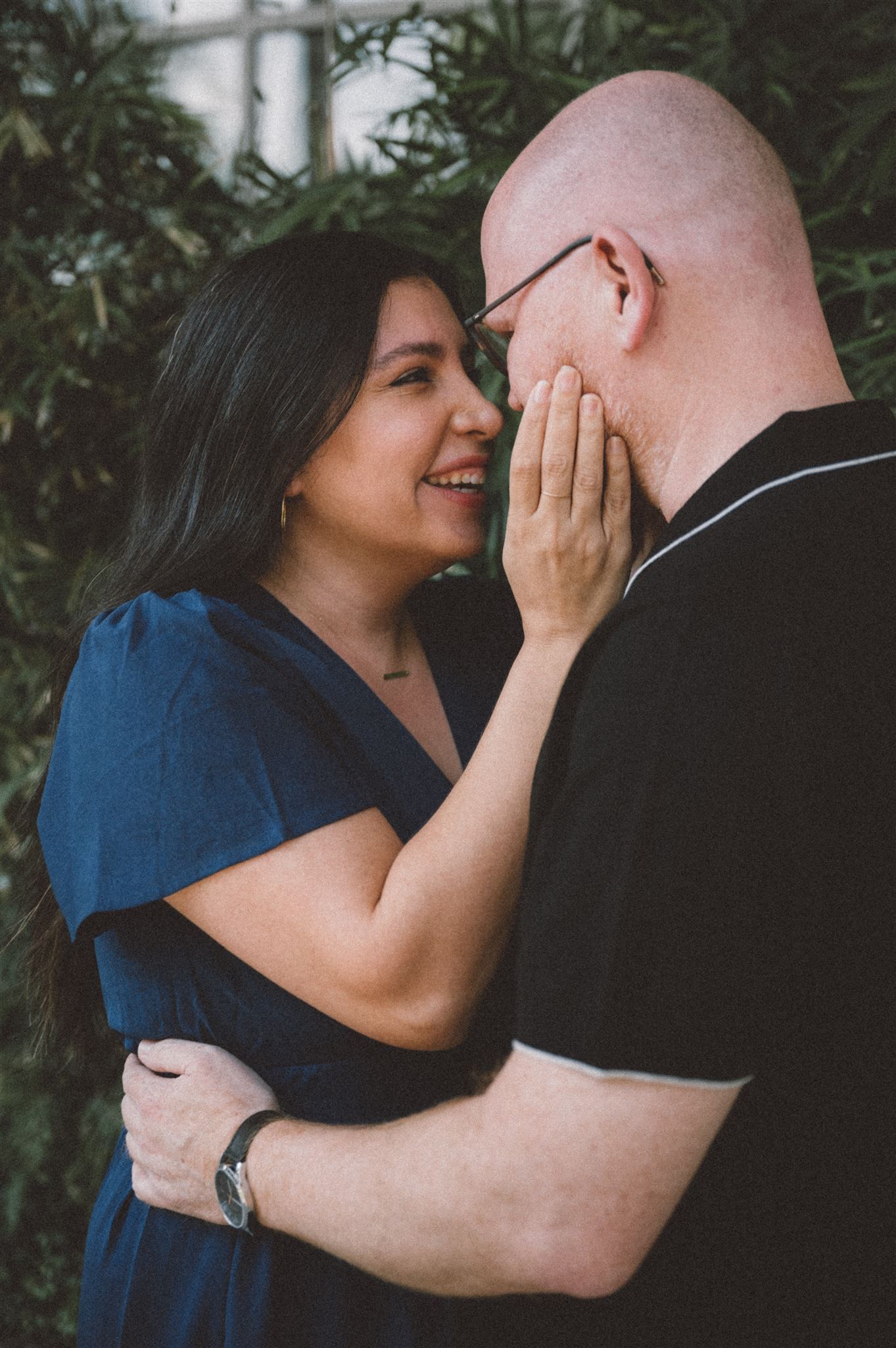 Tender embrace between couple wearing blue and black against green foliage.