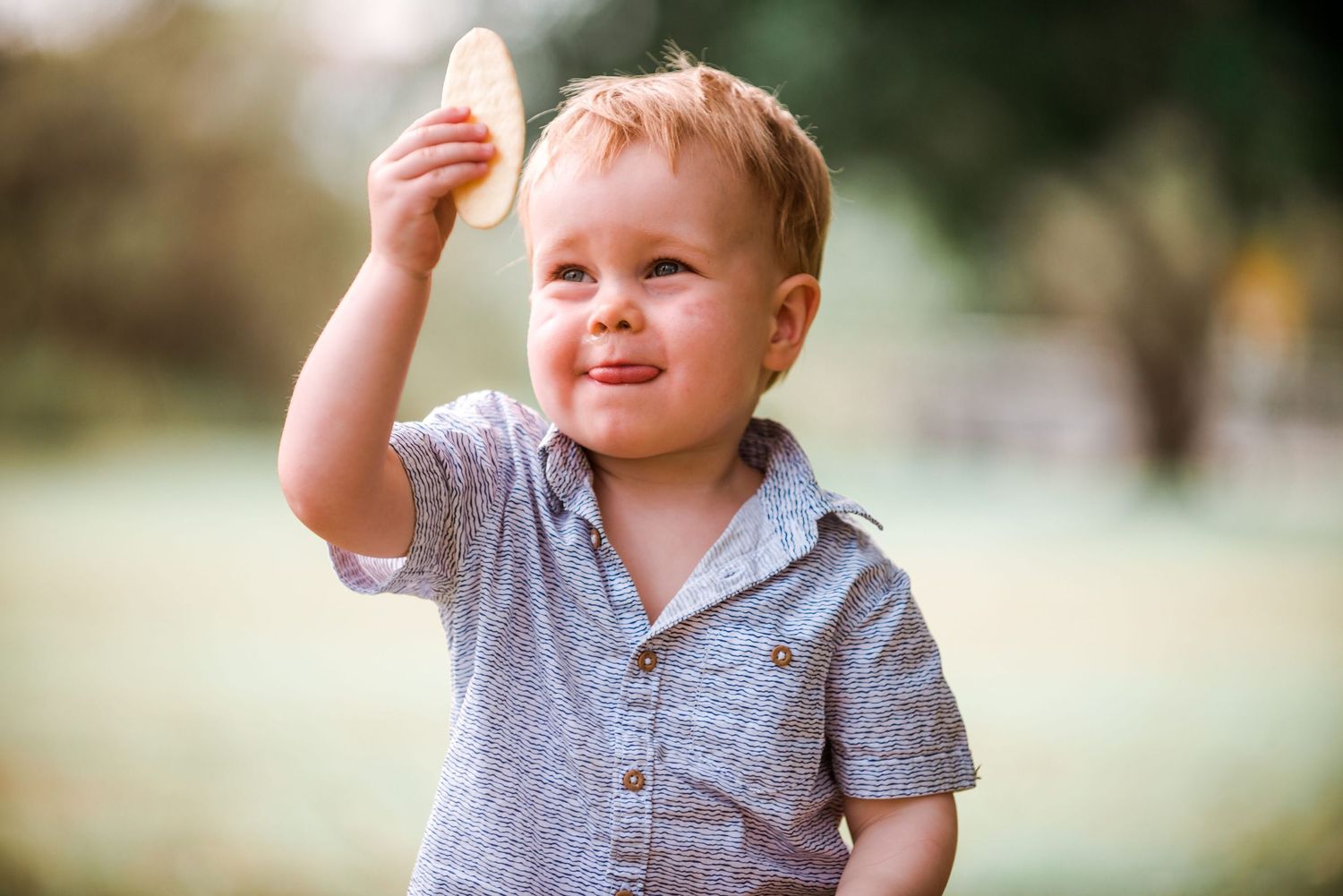 Toddler in grey shirt raising hand with joyful expression during outdoor photo session.