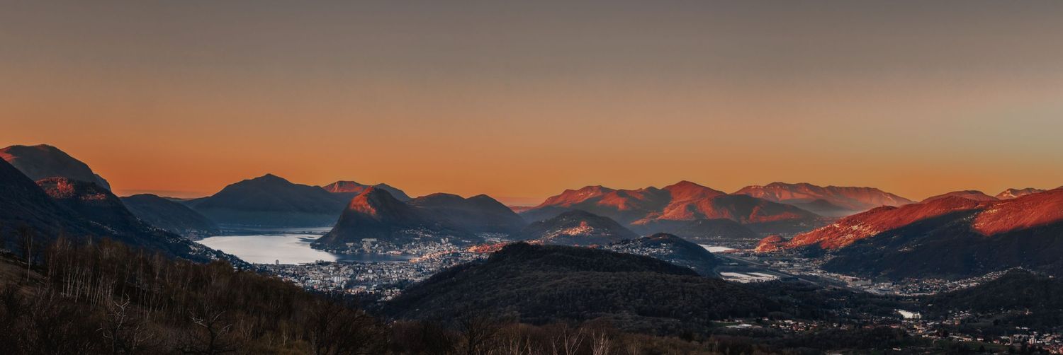 Panoramic mountain landscape at sunset with a lake and snow-capped peaks in the distance.