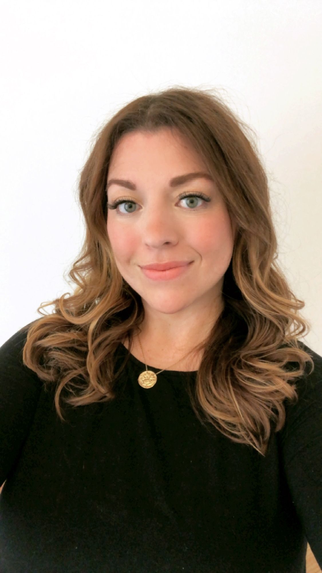 A woman with wavy brown hair wearing a black top and gold necklace smiles at the camera.