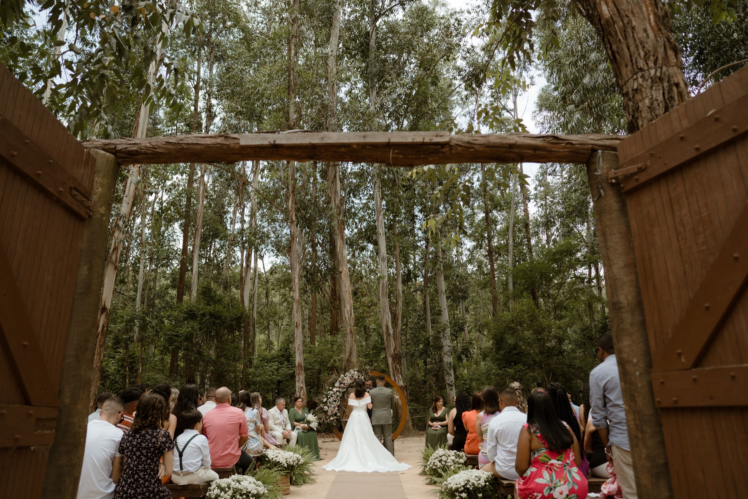Wide view of wedding ceremony through wooden doors in eucalyptus forest with seated guests.