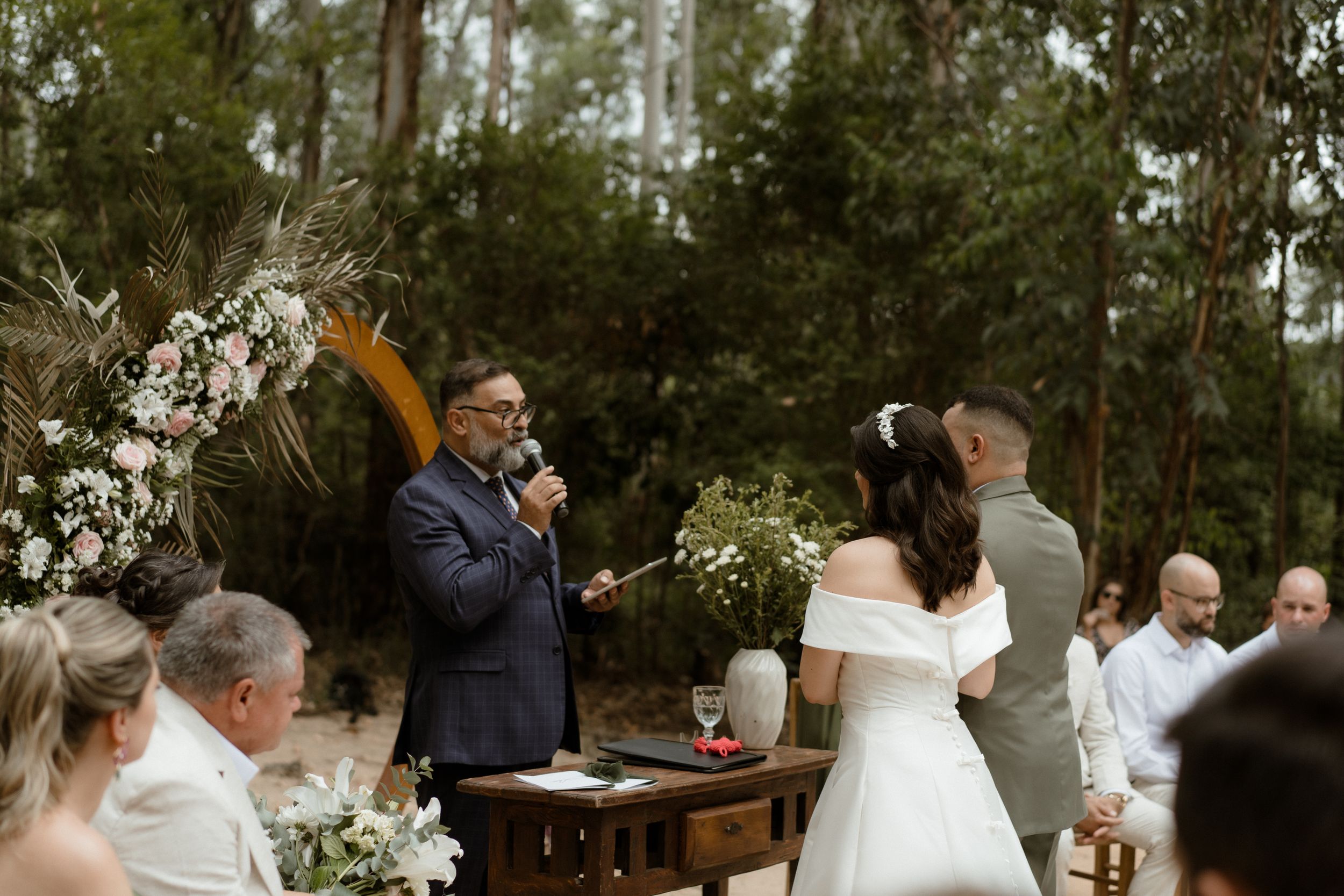 Wedding celebration moment with couple and officiant under tall forest canopy.