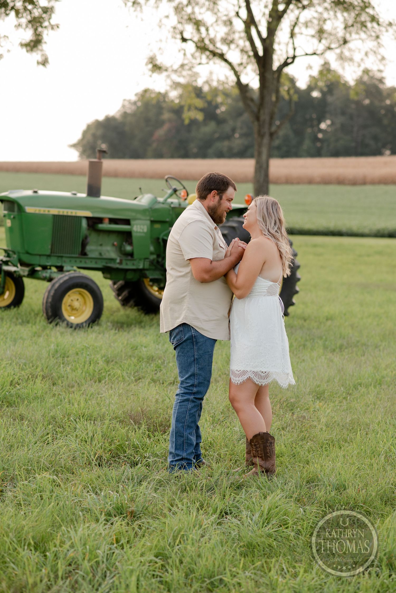 Couple in casual attire embraces near vintage green tractor in rural field at sunset.