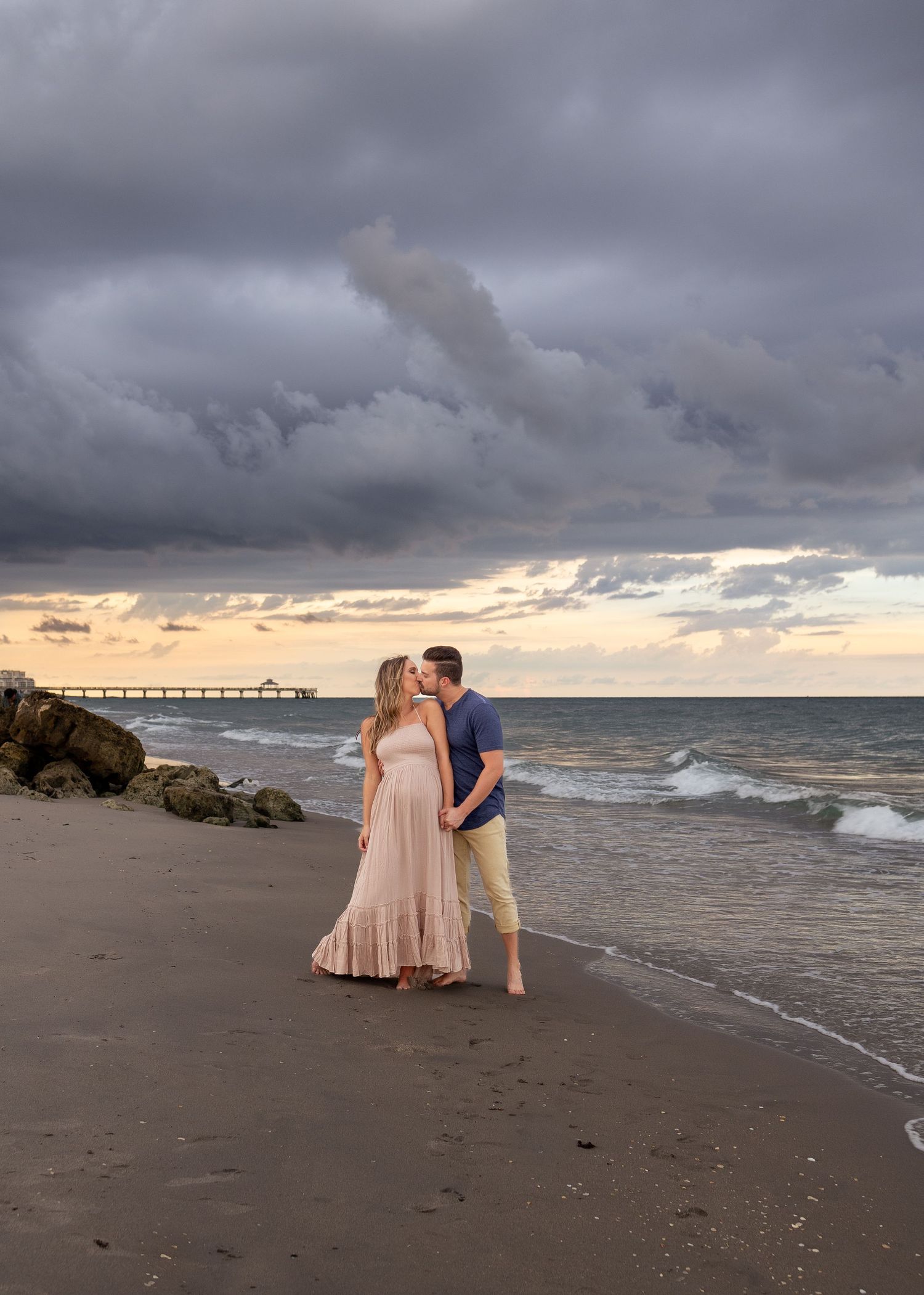 A romantic beach sunset scene with stormy clouds and a pier visible in the distance at the ocean.