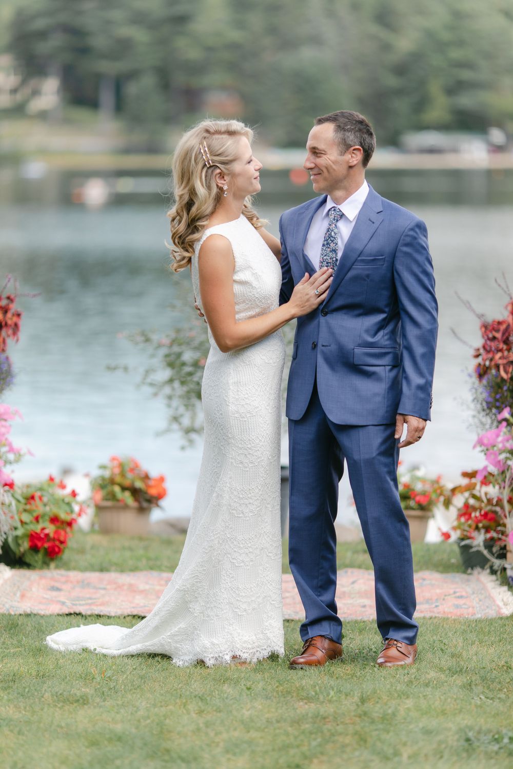 A wedding couple shares a joyful moment by a lake with colorful flowers in the foreground.