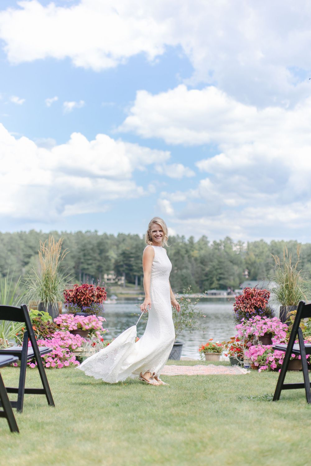 A scenic lakeside wedding ceremony setup with pink flowers and black chairs on a grassy area.