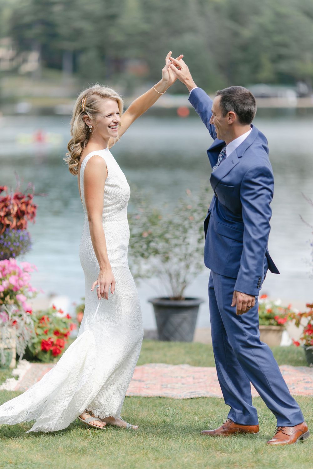 A bride and groom dance joyfully on their wedding day by a lake with flowers.