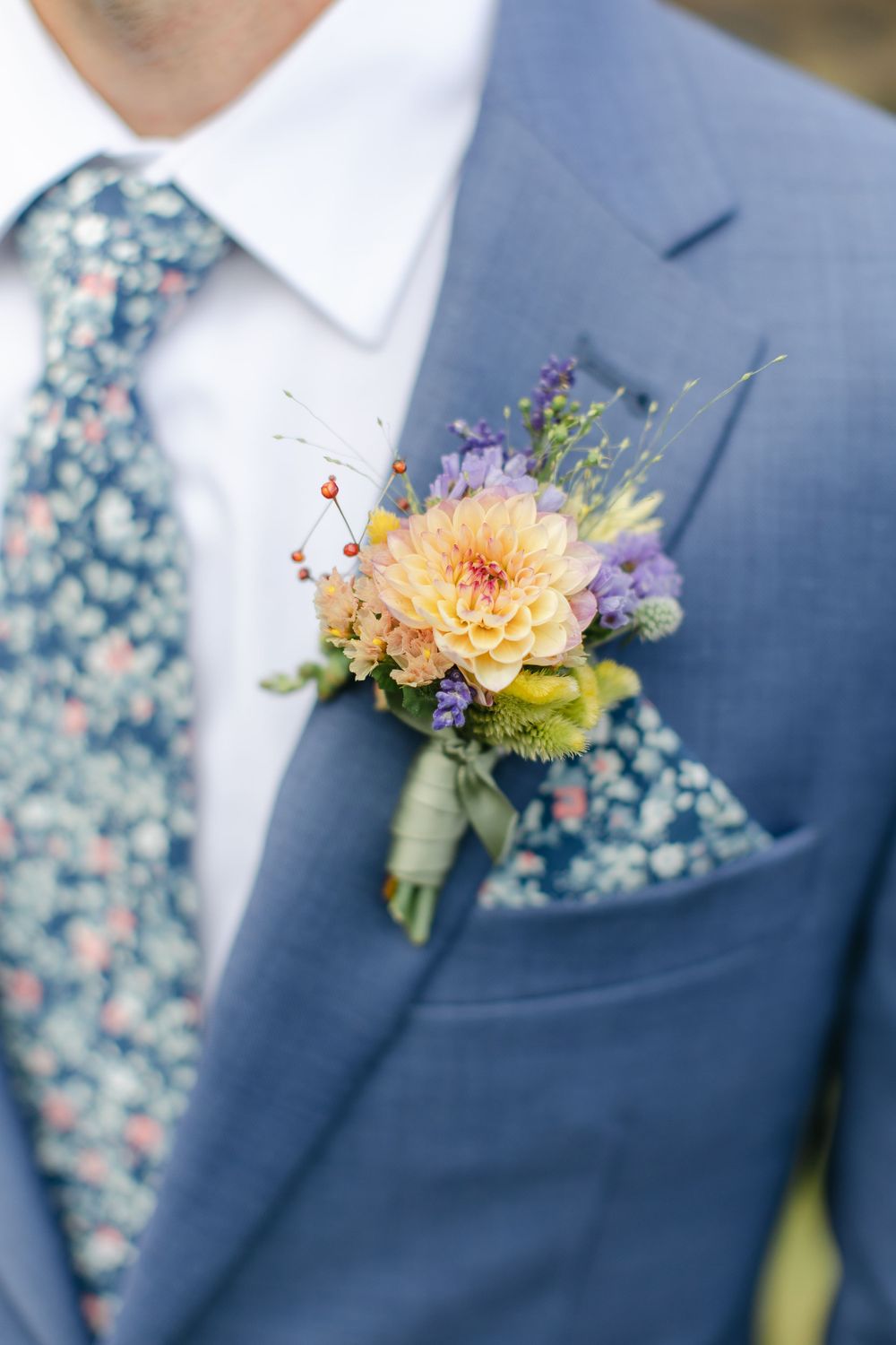 A detailed shot of a blue suit lapel with a peach dahlia and purple wildflower boutonniere.