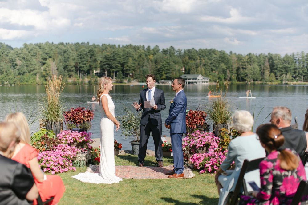 A lakeside wedding ceremony with pink floral decorations and pine trees in the background.