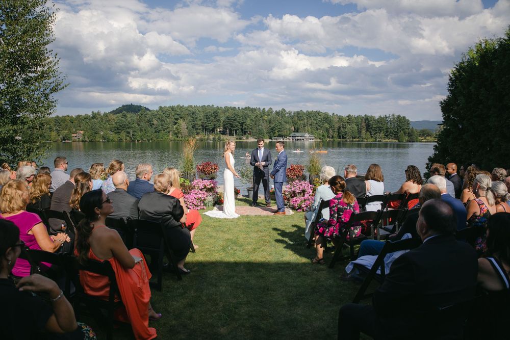 A wide view of an outdoor wedding ceremony by a lake with guests seated on both sides.