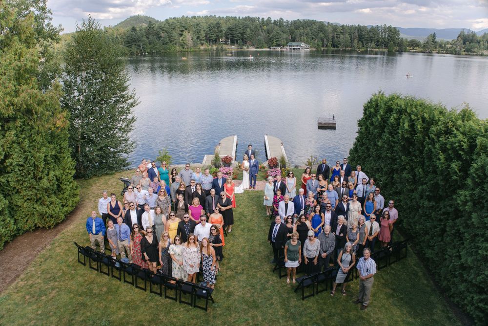 Aerial view of wedding ceremony guests seated in white chairs on a grassy lawn beside a serene lake.