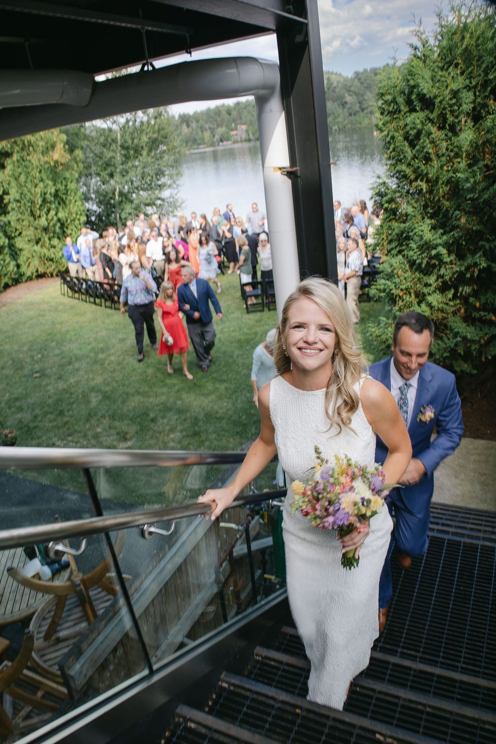 Wedding guests gather on lakeside lawn while couple ascends outdoor staircase.