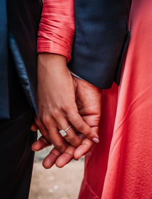 Close-up of two hands clasped together showing an engagement ring, with red dress fabric and navy suit visible.