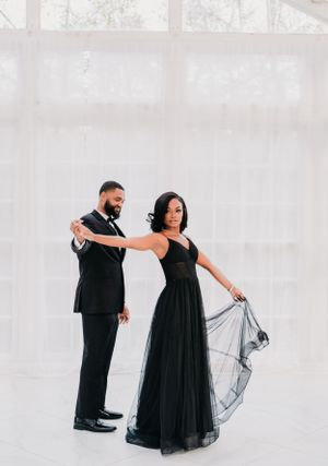 A couple in elegant black formal wear strike a graceful dance pose against a light background.