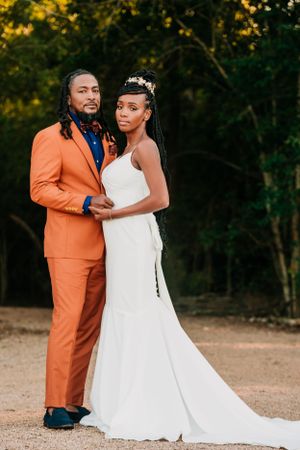 A couple poses together at their wedding, with the groom in a bright orange suit and bride in a flowing white gown.