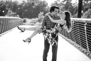 Black and white romantic photo of a couple embracing on a bridge, with the person wearing heels being held in a dip pose.