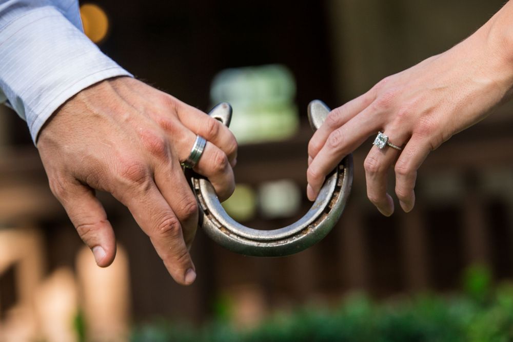 Two hands holding a horseshoe together, both wearing wedding rings with green foliage visible in background.