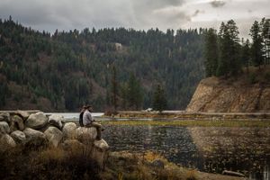 A person sits on rocky shoreline overlooking a serene mountain lake surrounded by evergreen forest on a cloudy day.