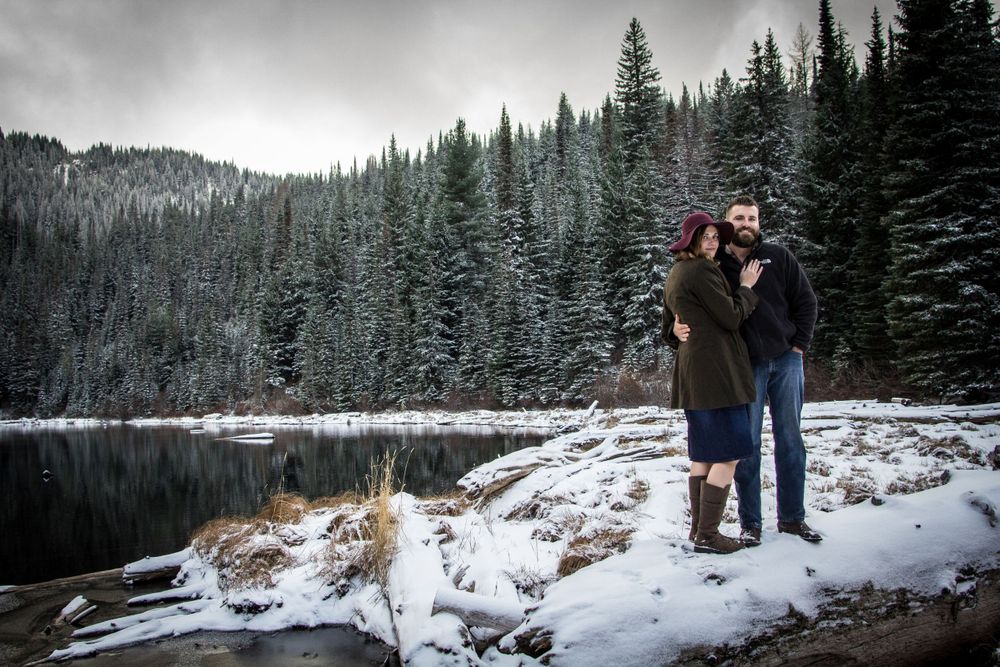 A couple stands by a snowy mountain lake surrounded by evergreen trees in a serene winter landscape.