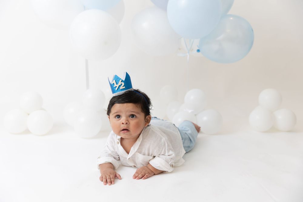 A baby in white lying on the floor with blue and white balloons and a tiny crown.