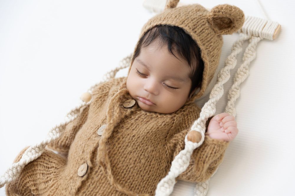 A sleeping newborn wearing a cozy brown knit outfit with bear ears lies on white blankets.