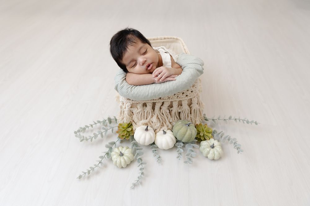 A sleeping newborn in white rests in a textured basket with decorative pumpkins and eucalyptus.