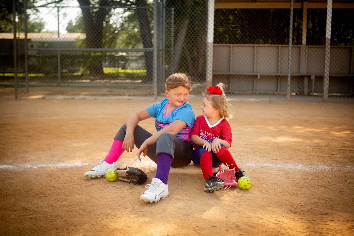 Two young softball players rest together on dusty baseball field.