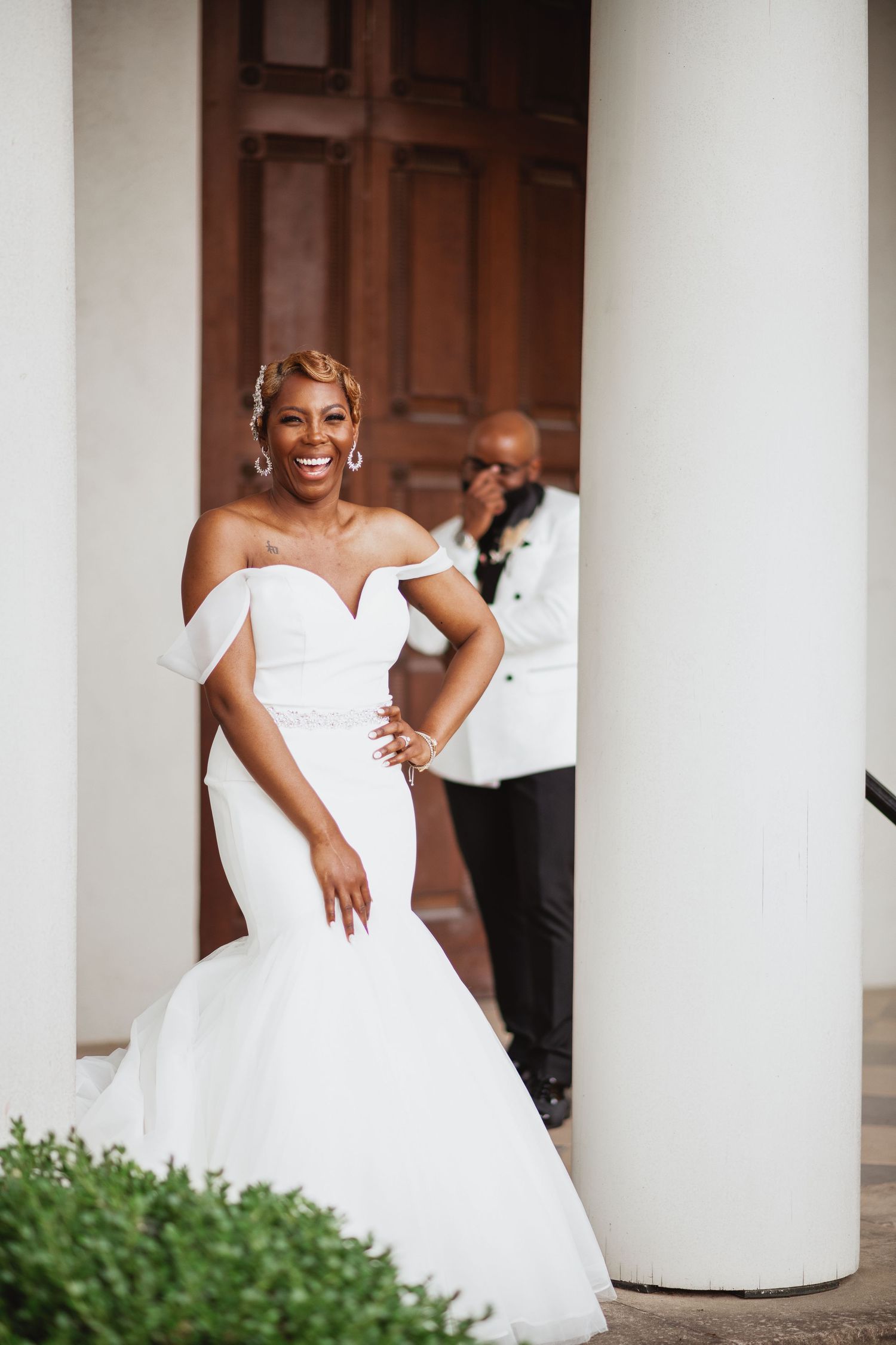 Radiant bride in off-shoulder mermaid gown poses between white columns while emotional groom reacts in background.