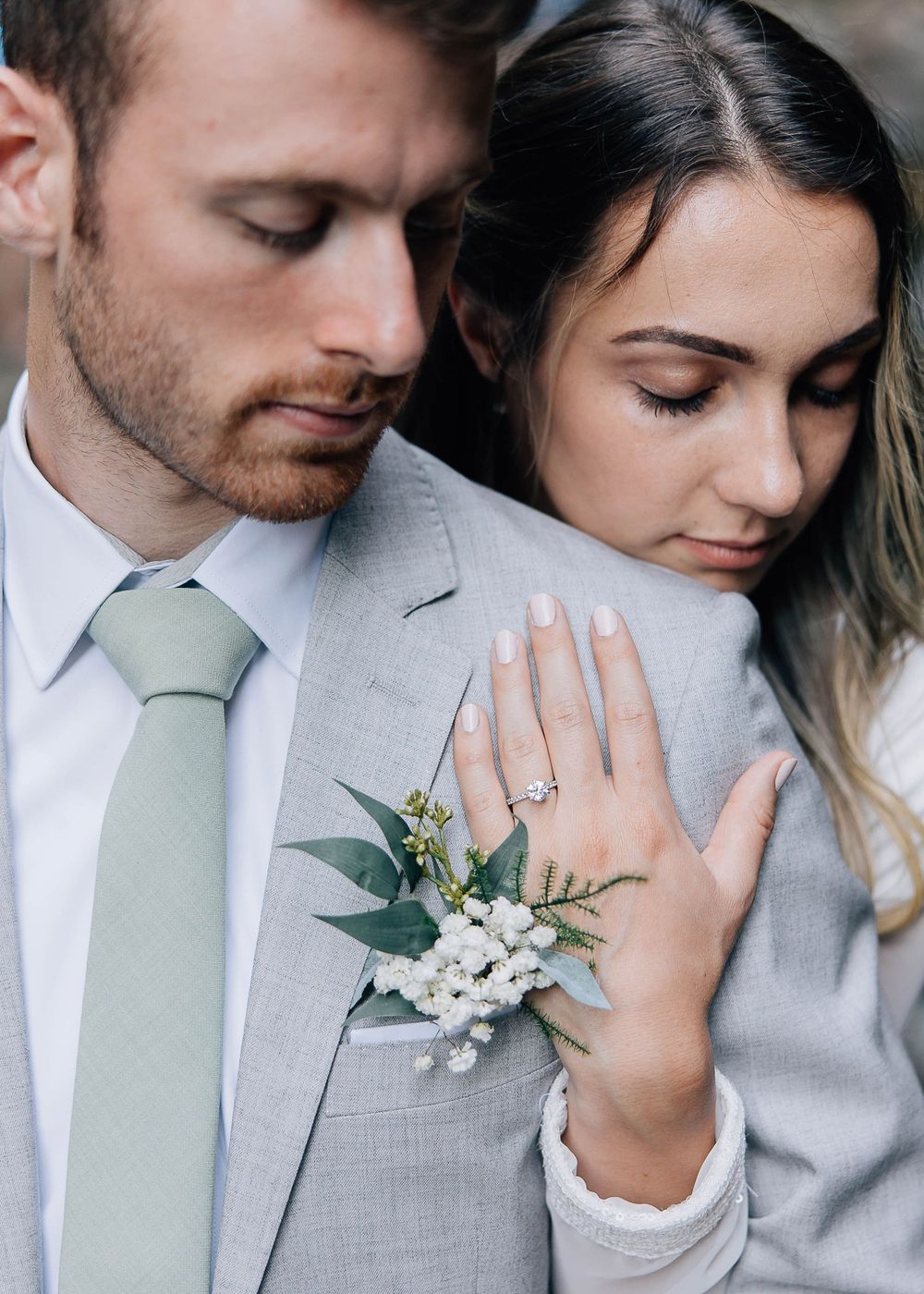 Close-up engagement photo showing a white floral boutonniere and wedding ring against a gray suit.