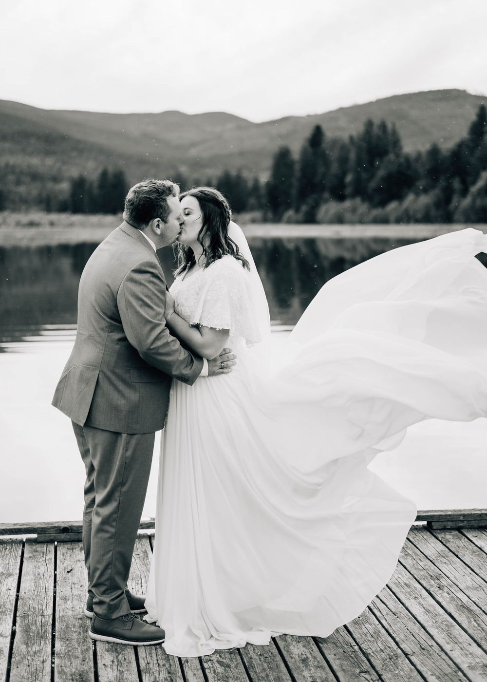 Romantic black and white wedding photo on a dock with flowing dress and mountain lake backdrop.