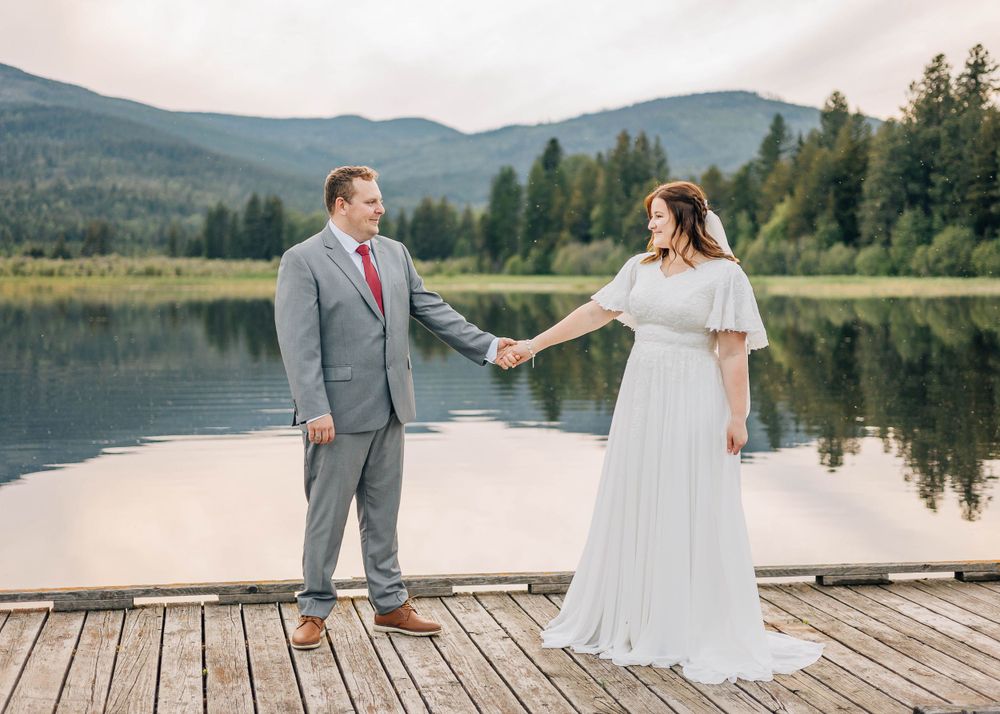 Newlyweds holding hands on wooden dock with mountain lake and forest scenery.