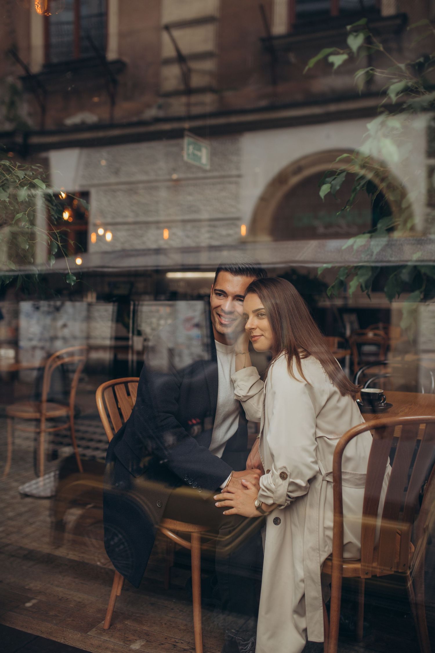 Couple sharing intimate moment seen through cafe window with city architecture in background.
