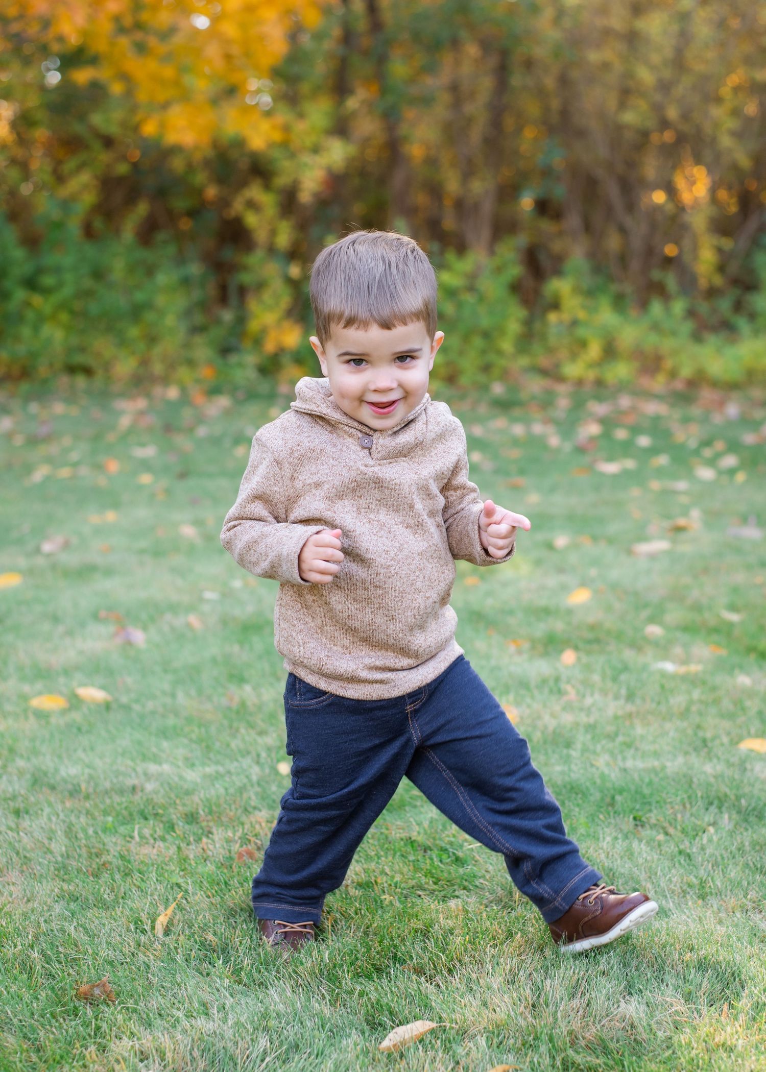 Young child in a beige hoodie plays on grass with autumn trees in the background.
