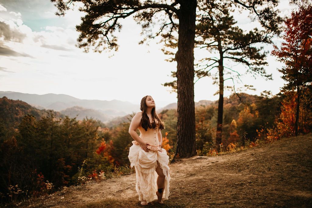 A scenic autumn mountain view with a solitary figure in a white dress sitting beneath towering pine trees.