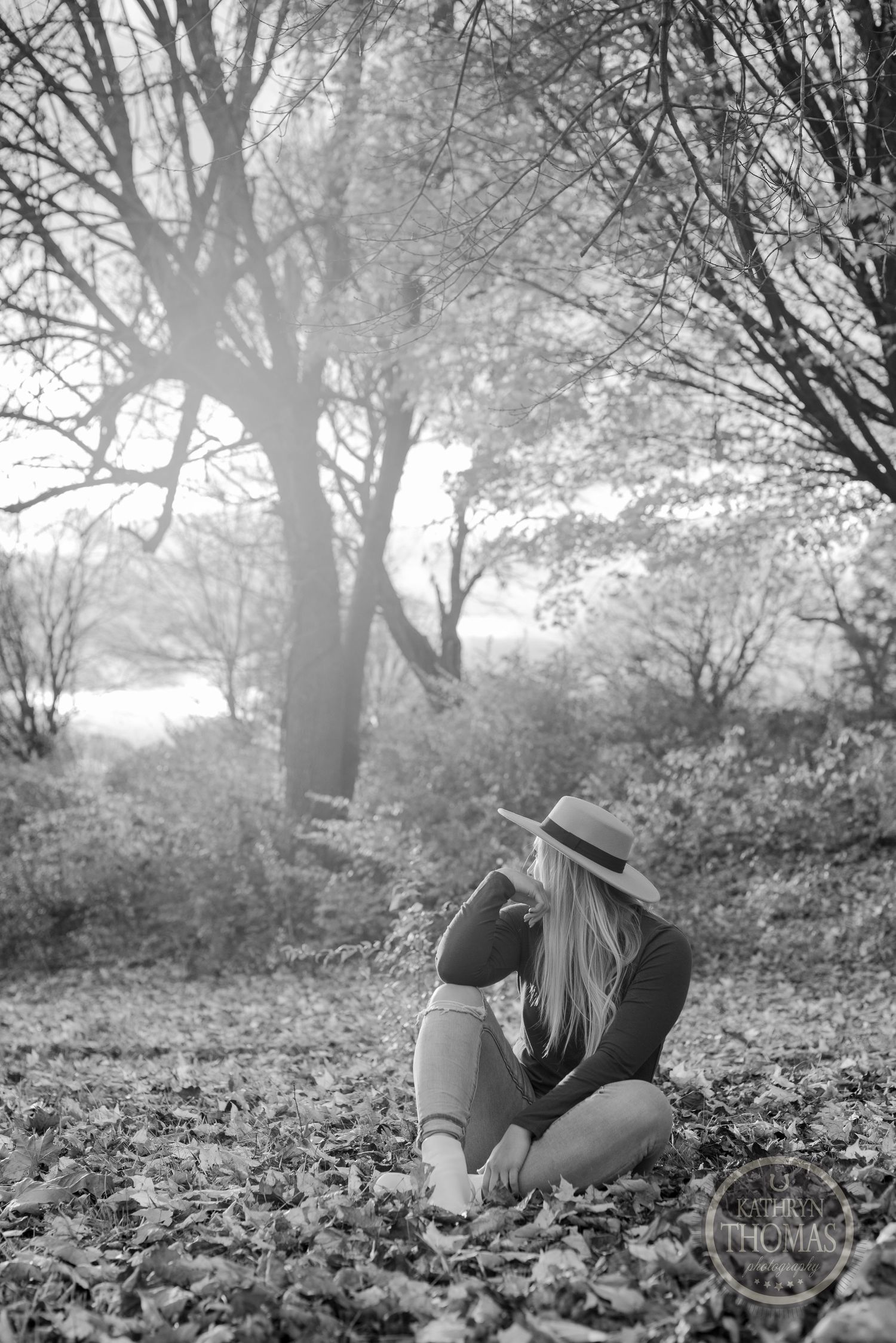 A figure in a wide brim hat sits on fallen autumn leaves in a black and white outdoor portrait.