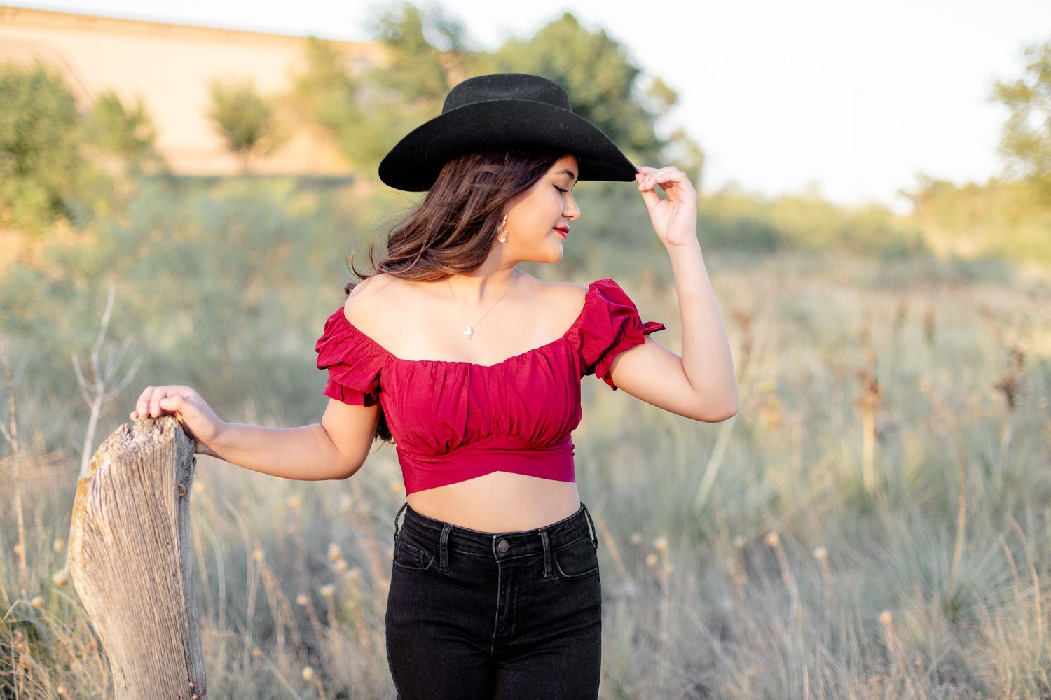 A woman in a red crop top and black hat poses in a rustic outdoor setting.