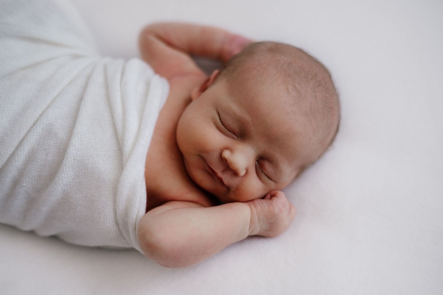 Sleeping newborn with slight smile wrapped in white fabric against light backdrop.