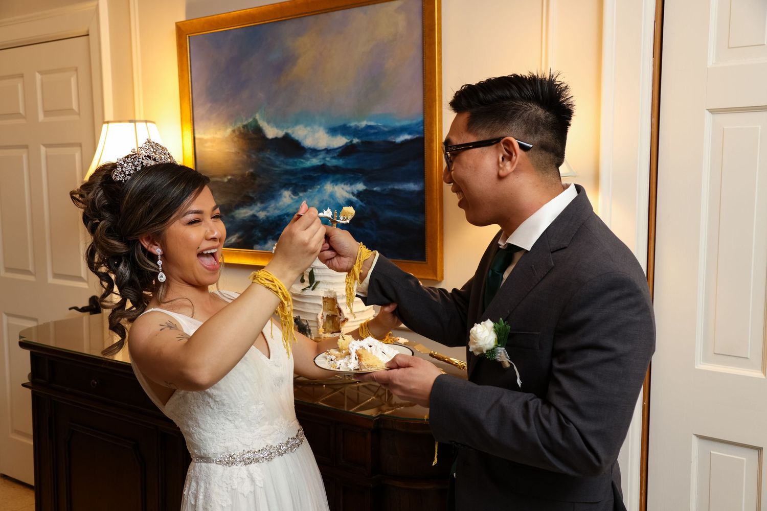 A joyful moment of cake feeding during a wedding reception with a seascape painting in the background.