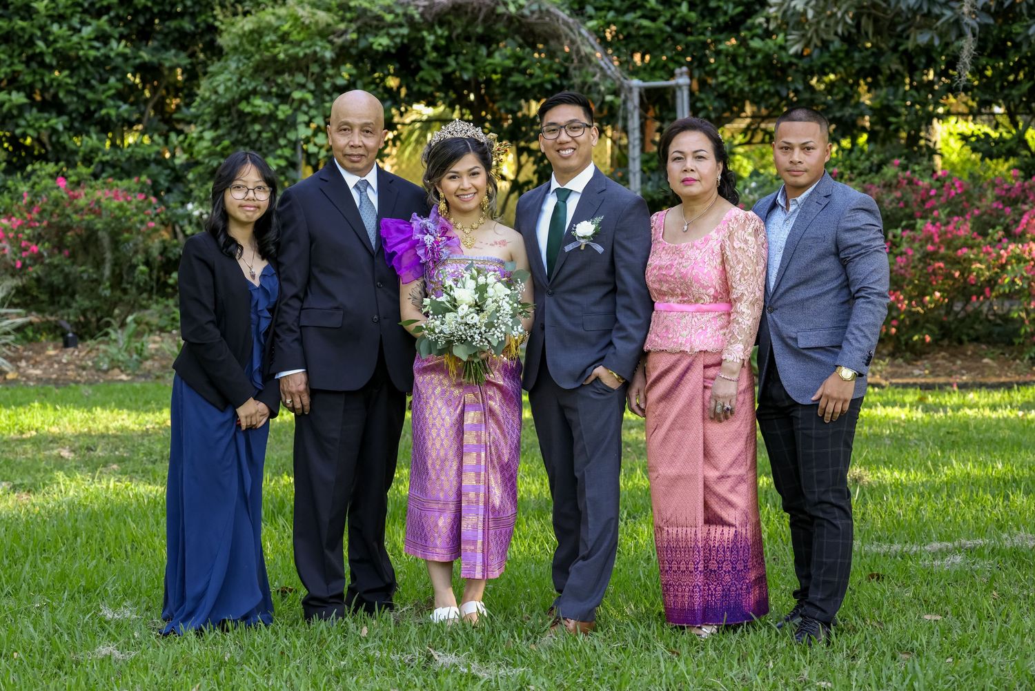 A formal group photo of wedding party members in traditional Asian attire standing in a garden setting.